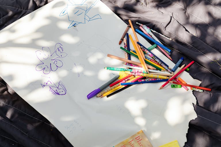 A collection of colored pencils and drawings on white paper under dappled sunlight, fostering creativity.