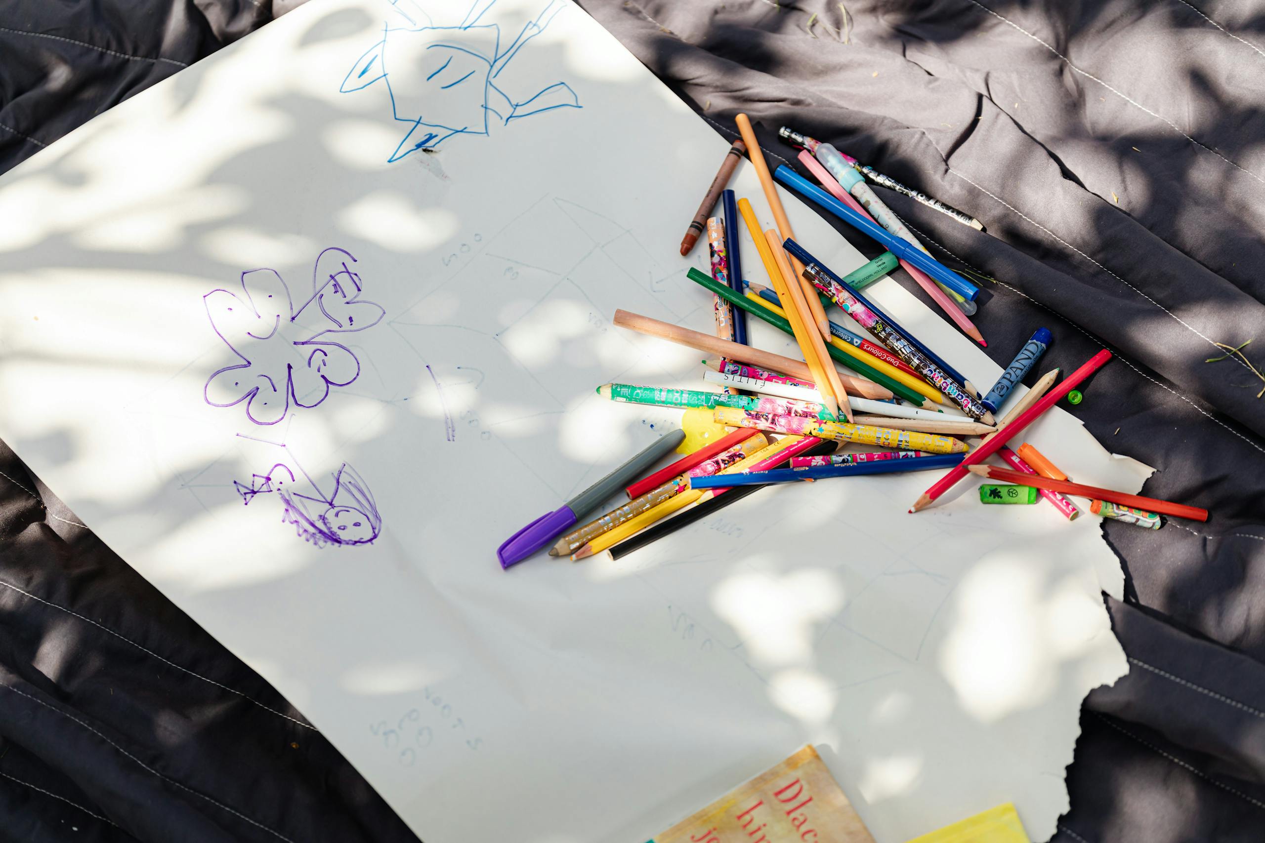 A collection of colored pencils and drawings on white paper under dappled sunlight, fostering creativity.